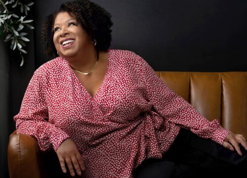 Gina relaxes on dark tan leather couch with a black wall behind her and an olive tree.
