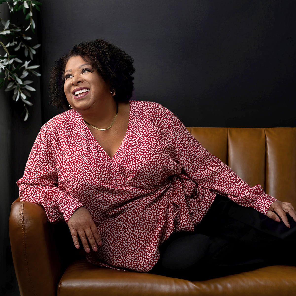 Gina relaxes on dark tan leather couch with a black wall behind her and an olive tree.