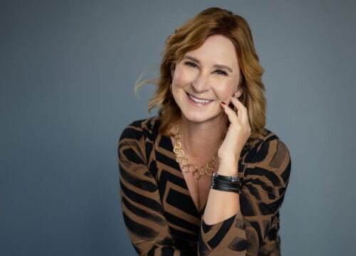 Woman holding her hand near face. In studio with grey background. Wearing brown and black striped wrap dress. Reddish brown hair.