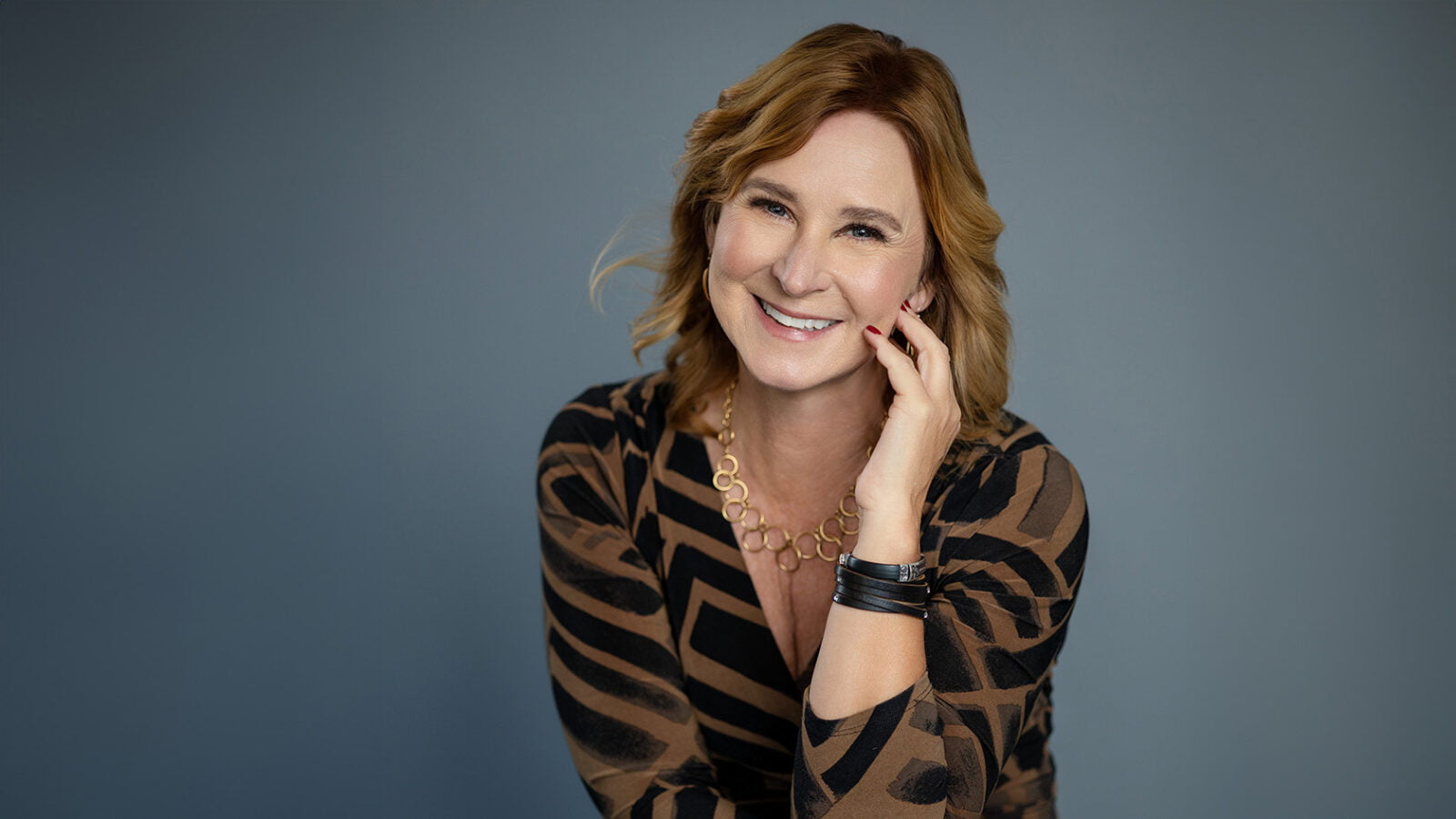 Woman holding her hand near face. In studio with grey background. Wearing brown and black striped wrap dress. Reddish brown hair.