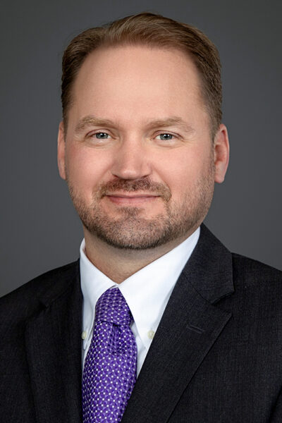 Bearded man in black suit with a purple tie and white shirt.