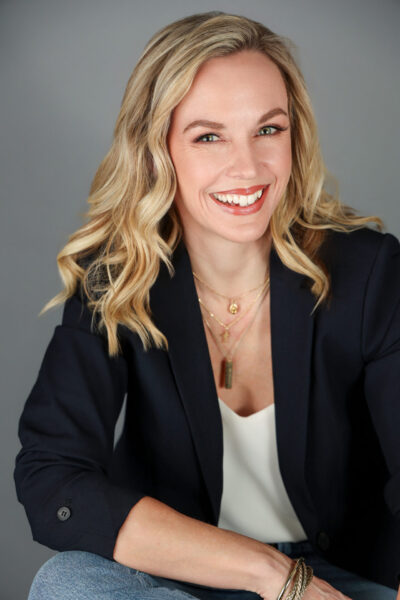 Blonde with a large smile and curls smiles and leans forward. She has a black blazer, cream top and jeans on. - Professional Columbus Photographer - Desired Focus Photography